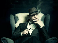 A private detective smokes a cigarello in an armchair. He is wearing a suit and a hat, and is holding a magnifying glass in his right hand.